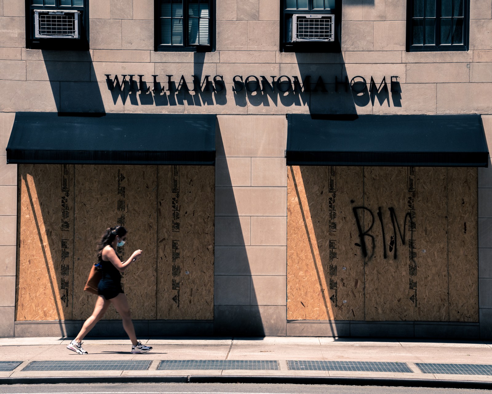 A pedestrian crosses geometric shadows in front of a boarded storefront.