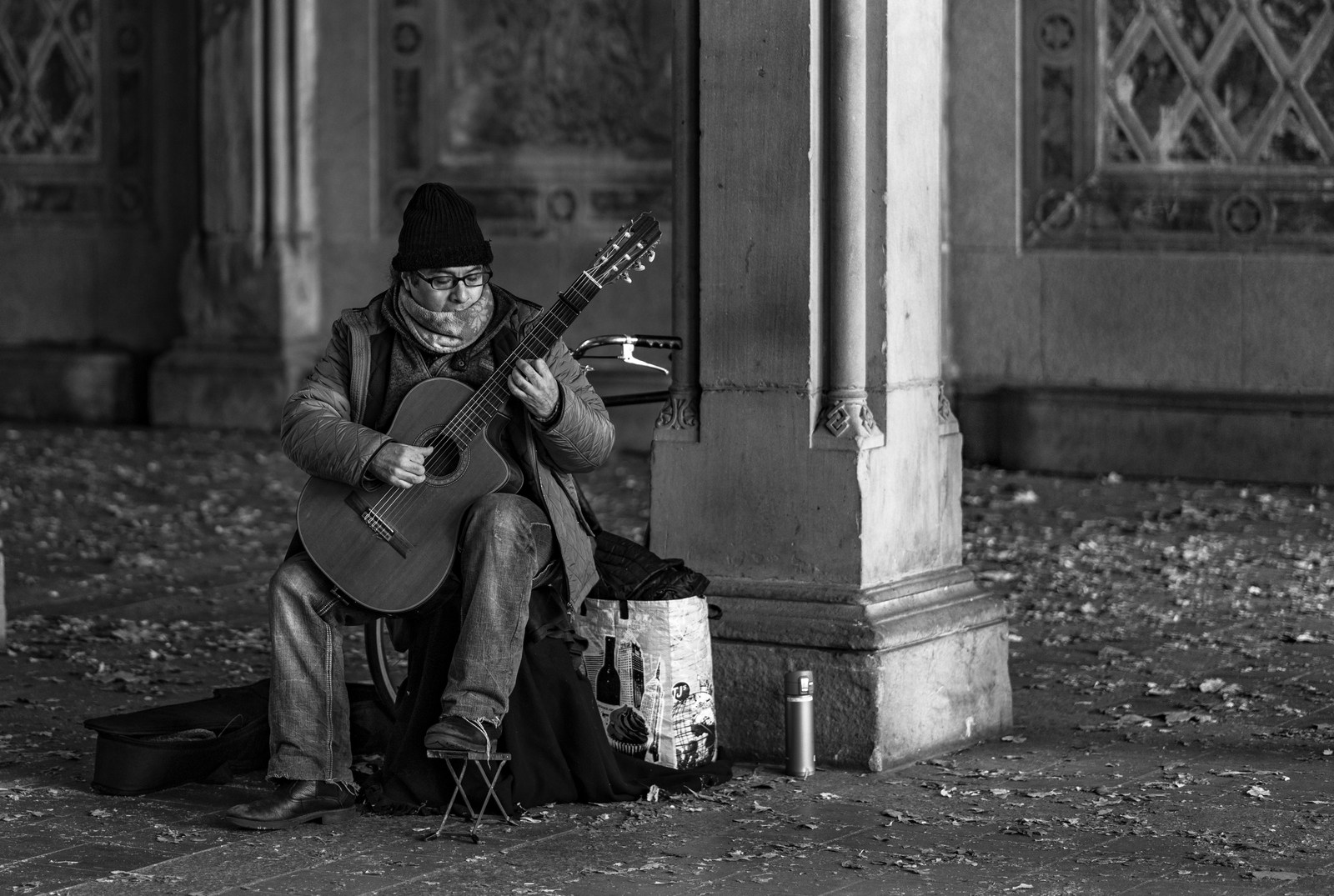 A seated guitarist plays beside a stone pillar and scattered fallen leaves in black and white.
