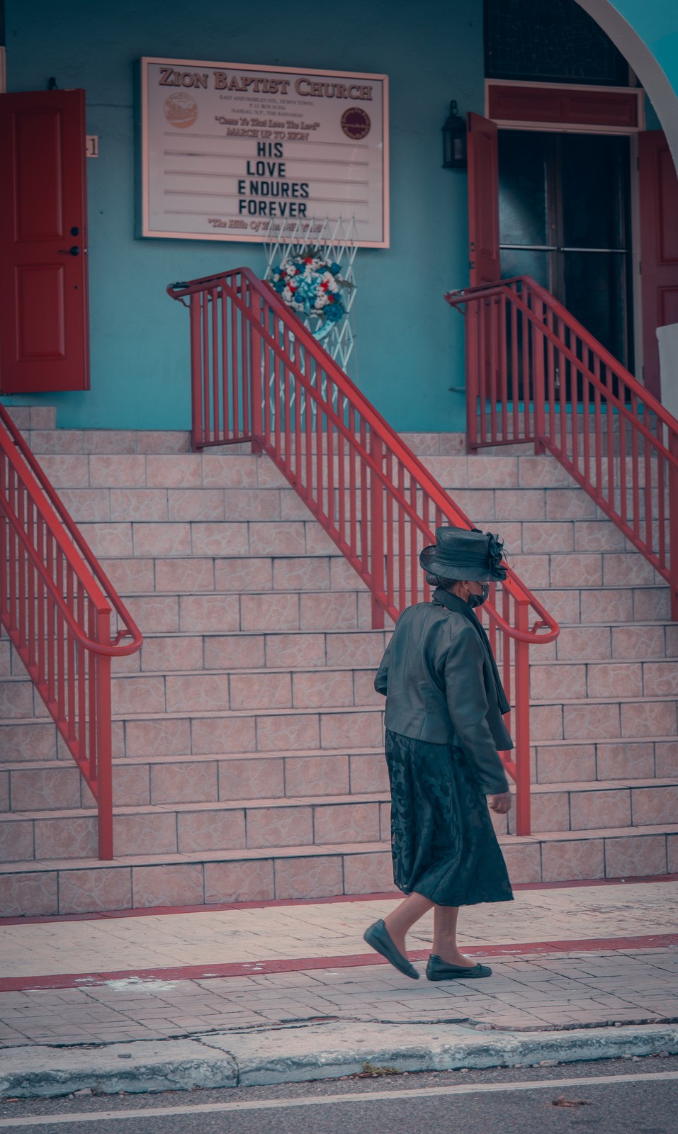 A woman in a dark hat and coat walks below red stair rails against a turquoise church facade.