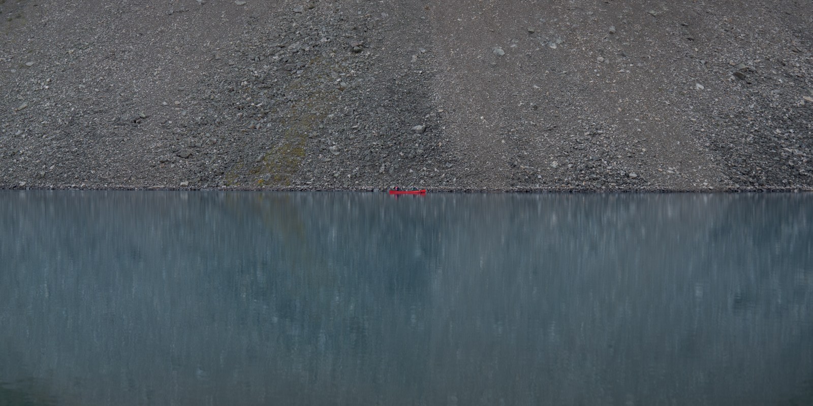 A tiny red canoe divides a grey scree slope from its blue reflection.
