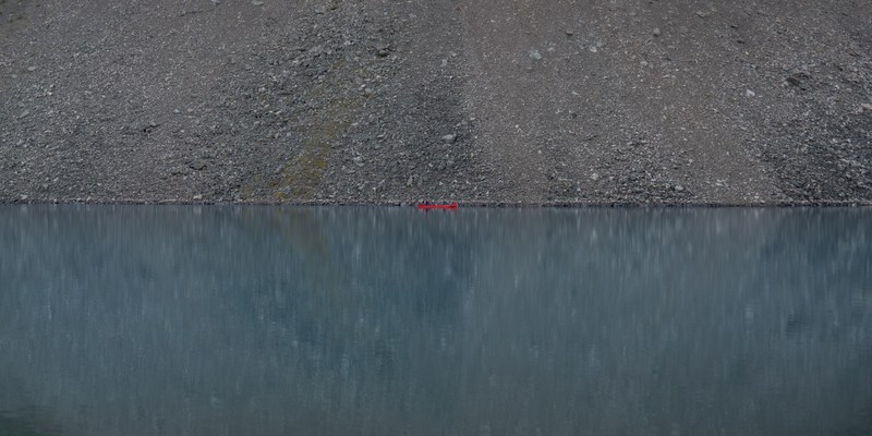 A tiny red canoe divides a grey scree slope from its blue reflection.