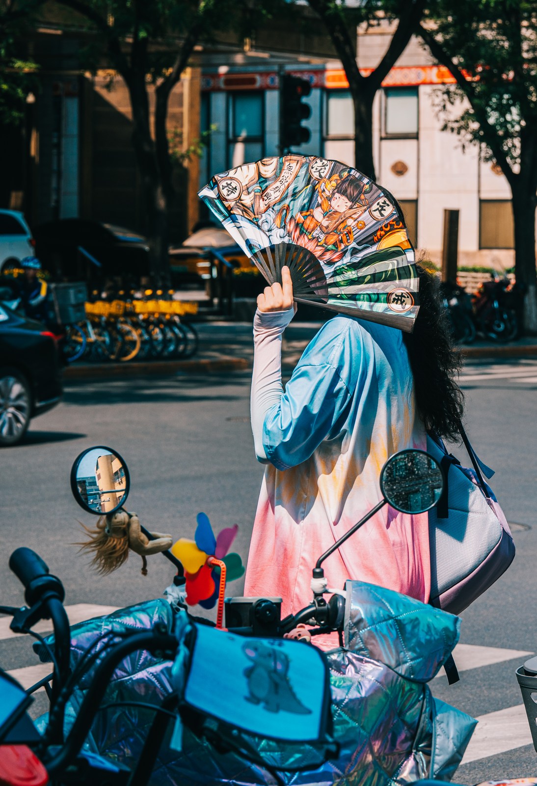 A person holds an illustrated fan over their face beside a decorated scooter.