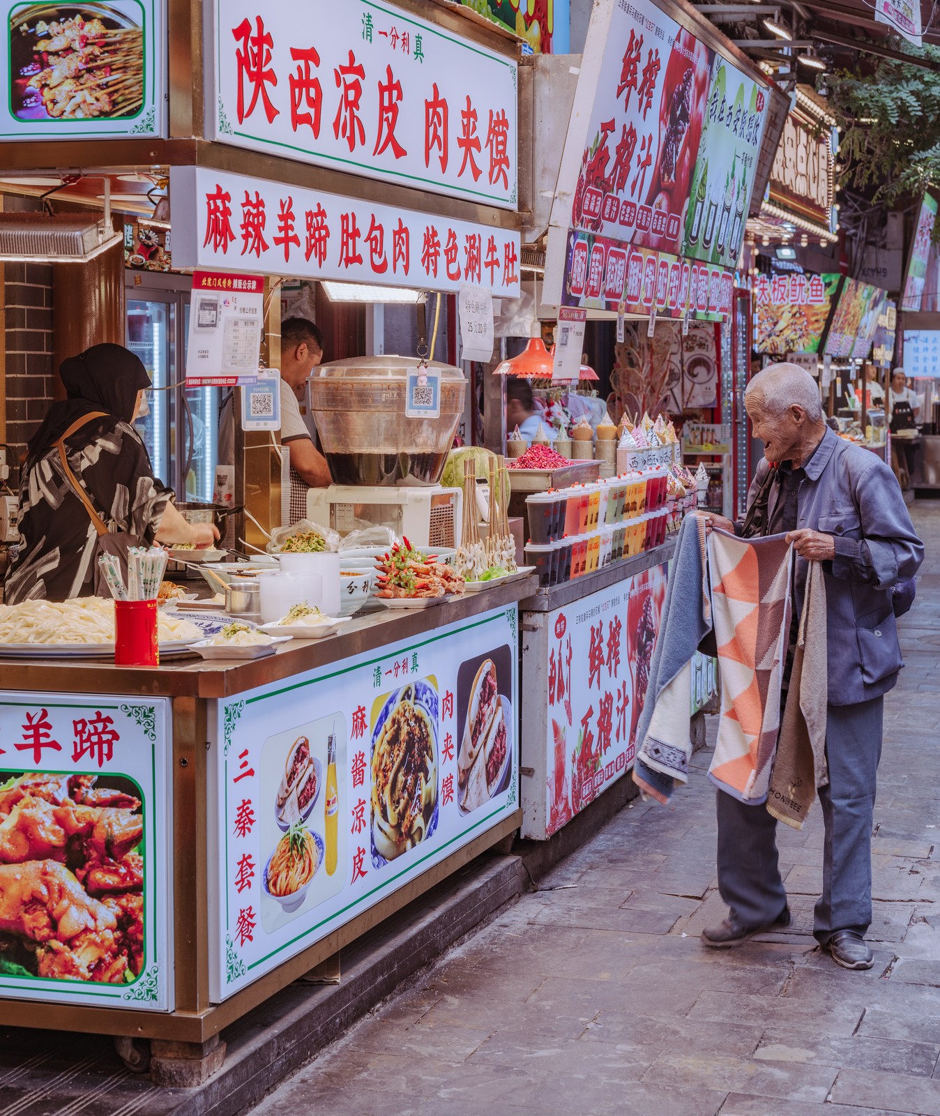 A man holds patterned fabric beside a row of food counters and signs.
