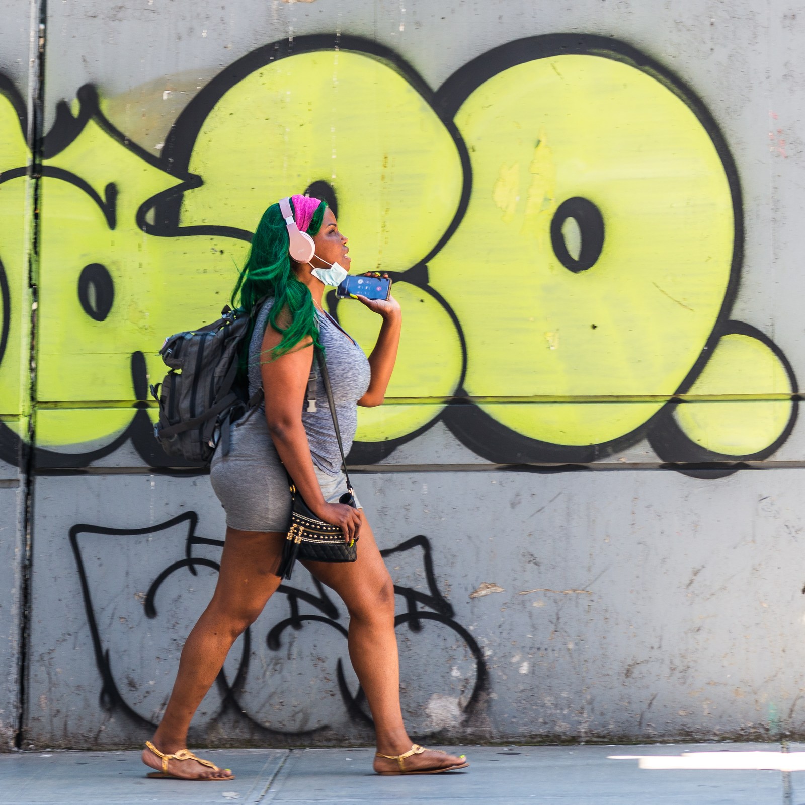 A pedestrian with green hair and pink headphones strides past yellow graffiti.
