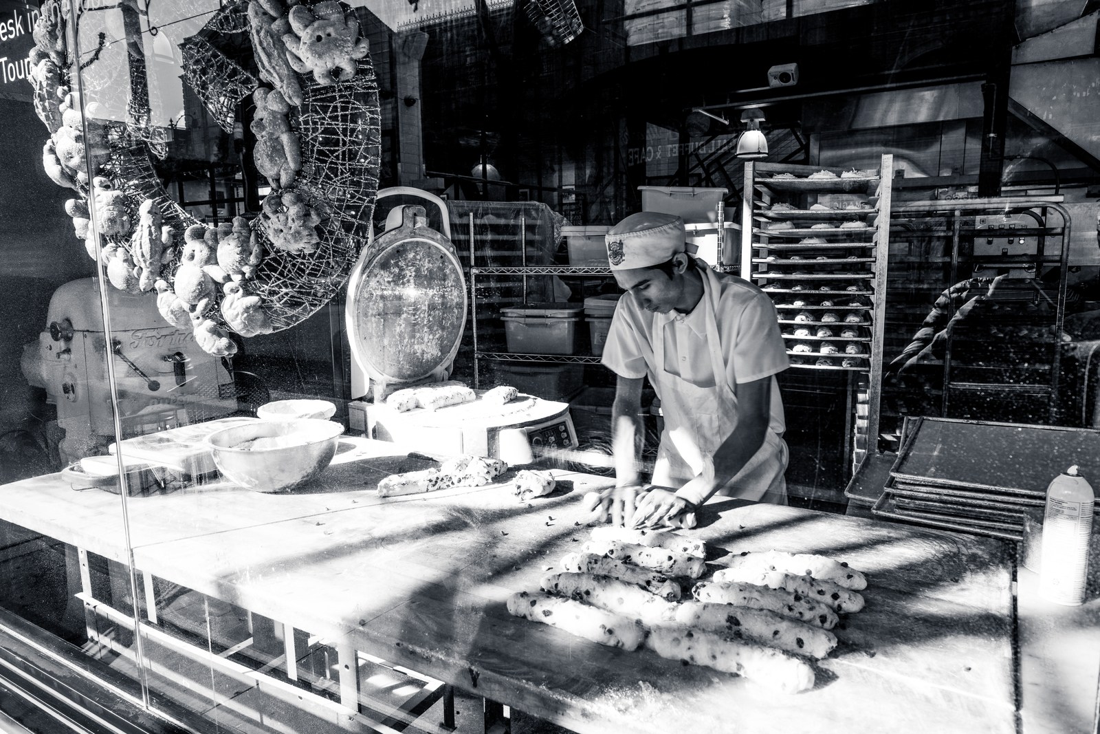 A baker works with dough behind reflective glass in a monochrome shop interior.