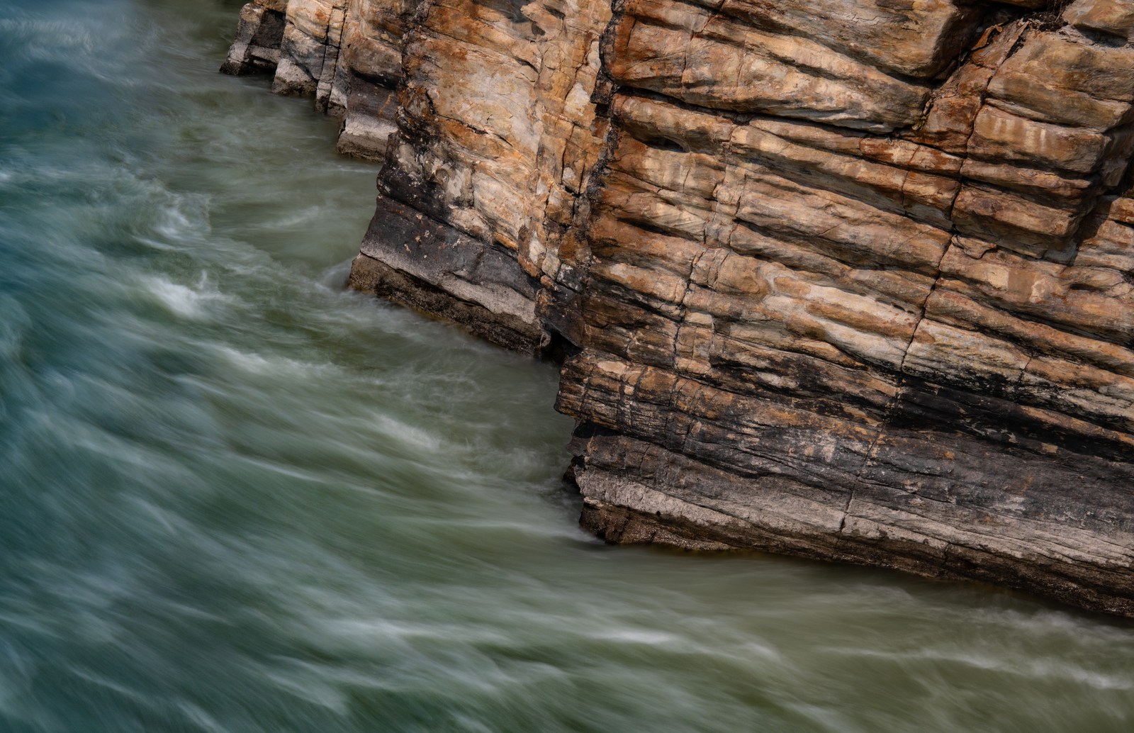 Horizontal ochre rock layers meet green water flowing around the base of a cliff.