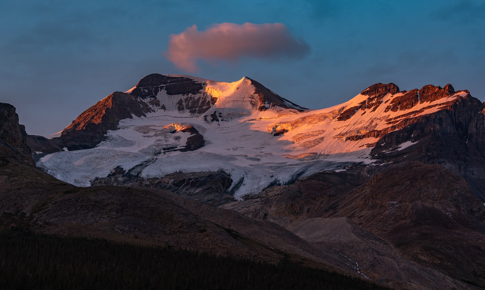Warm light touches snow and a small cloud above a dark mountain face.