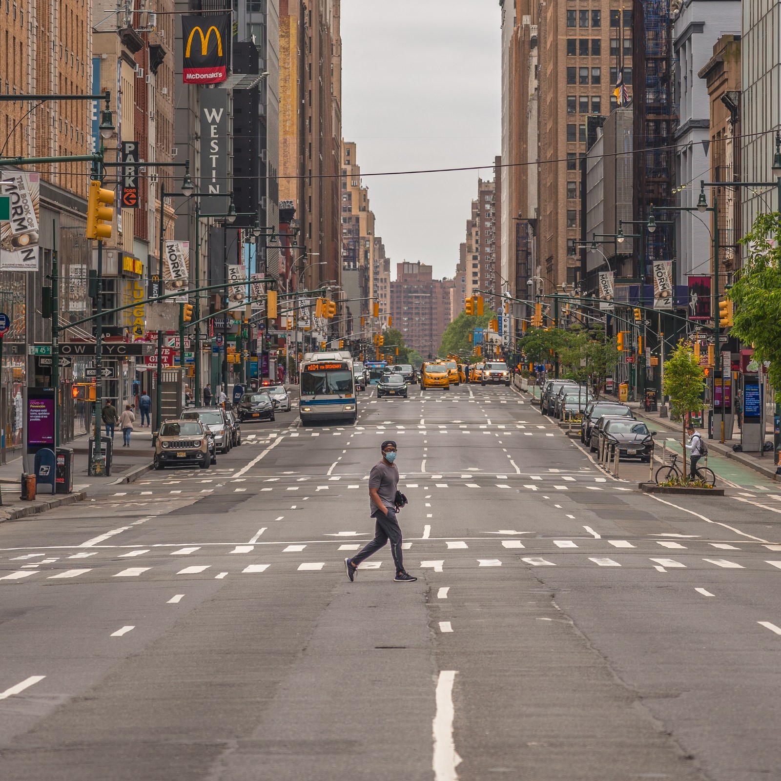 A lone pedestrian crosses a broad avenue between rows of buildings and distant traffic.