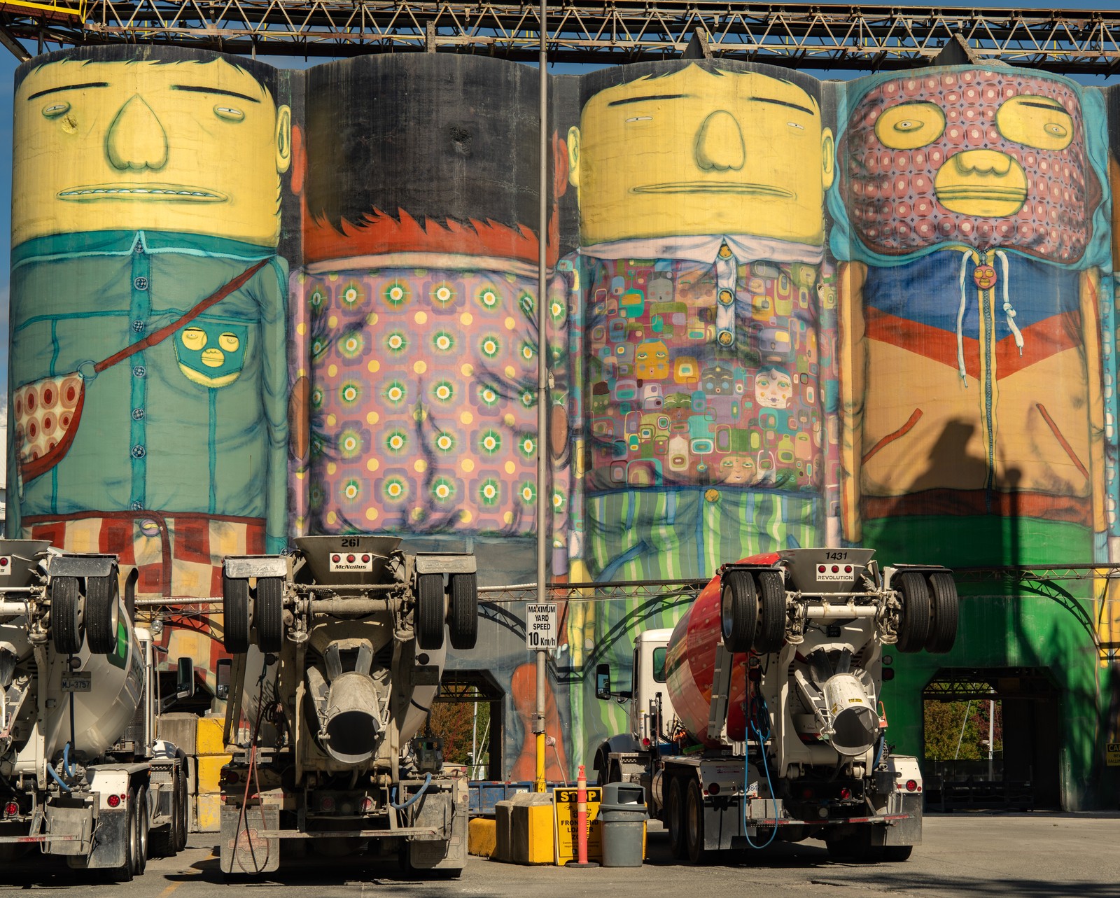 Colorful figures painted on industrial silos tower over a row of concrete mixer trucks.
