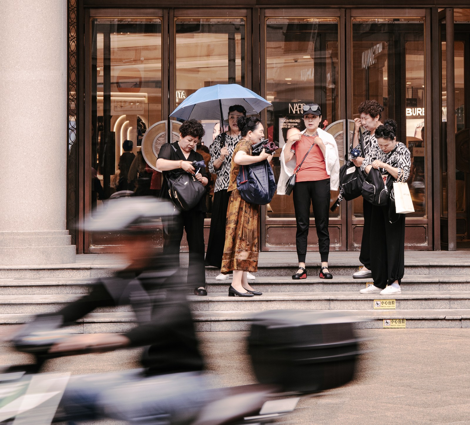 A group pauses on shop steps while a scooter crosses the foreground in a blur.