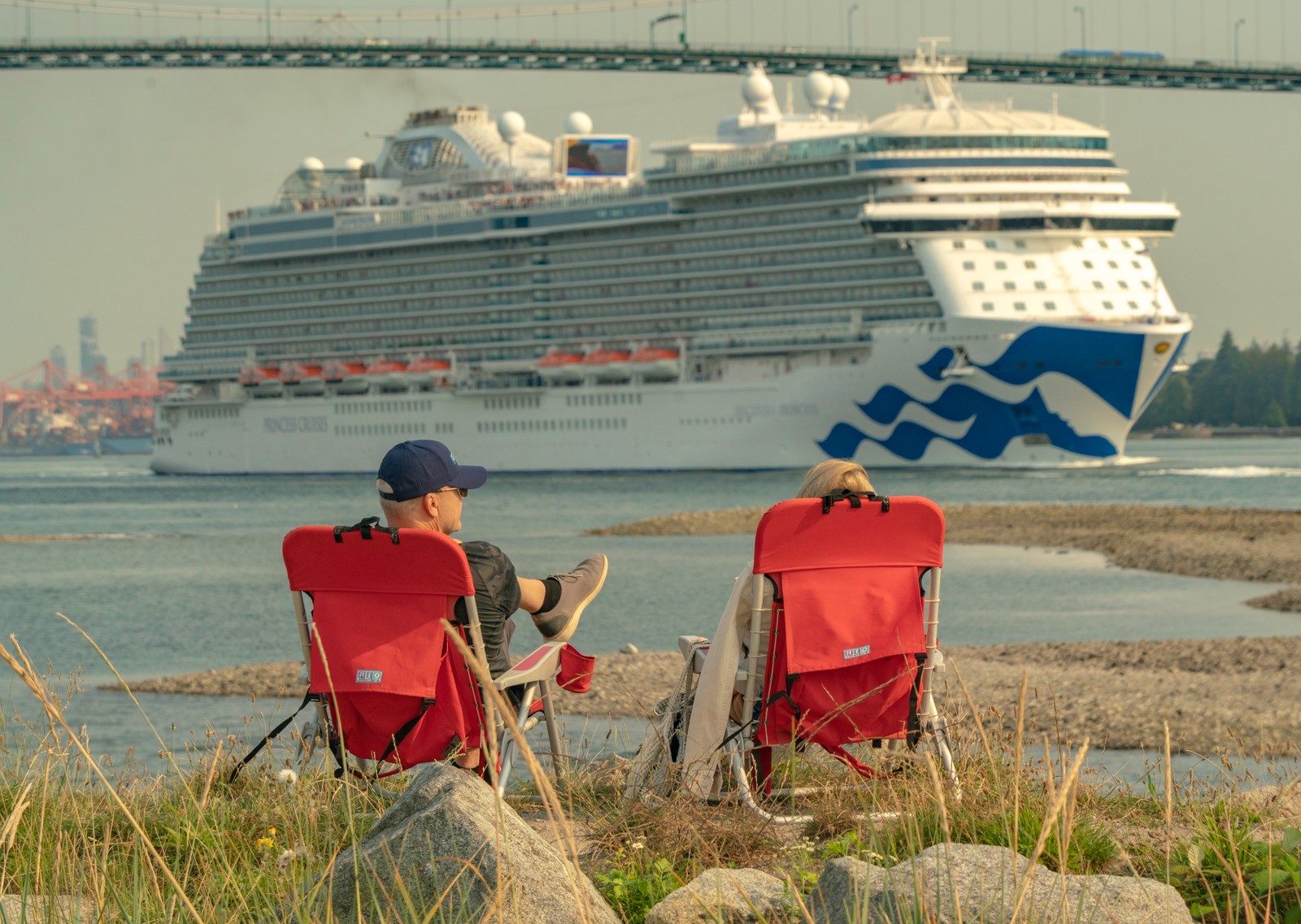 Two people in red folding chairs watch a large cruise ship pass beneath a bridge.