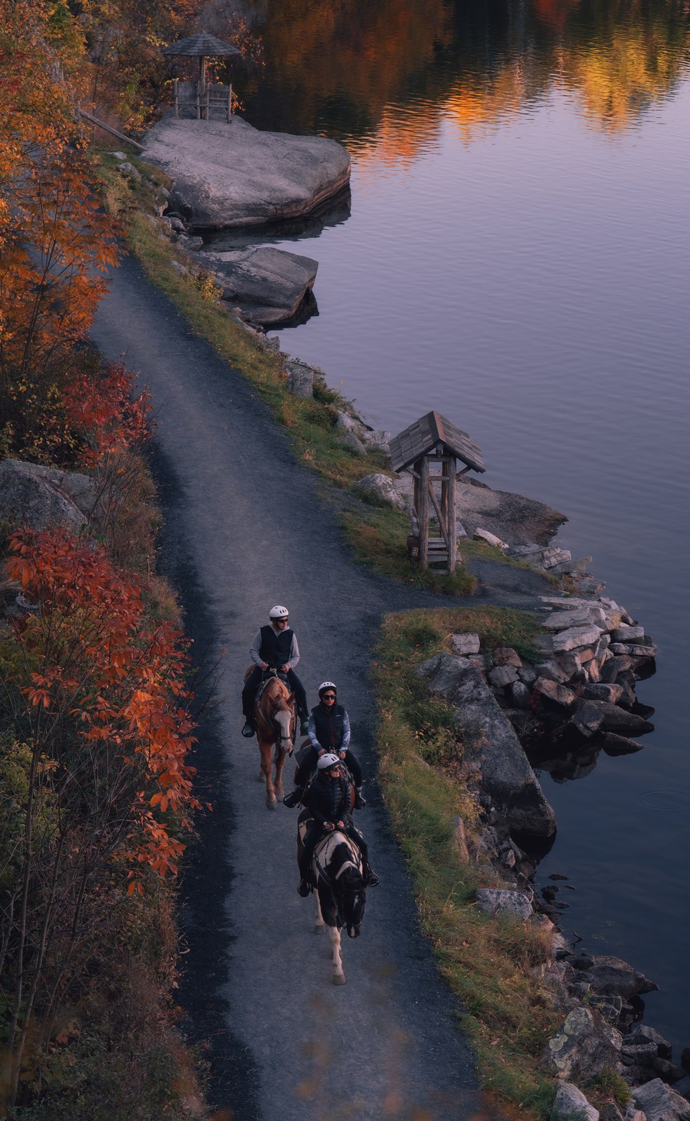 Three riders follow a narrow autumn path beside still water.