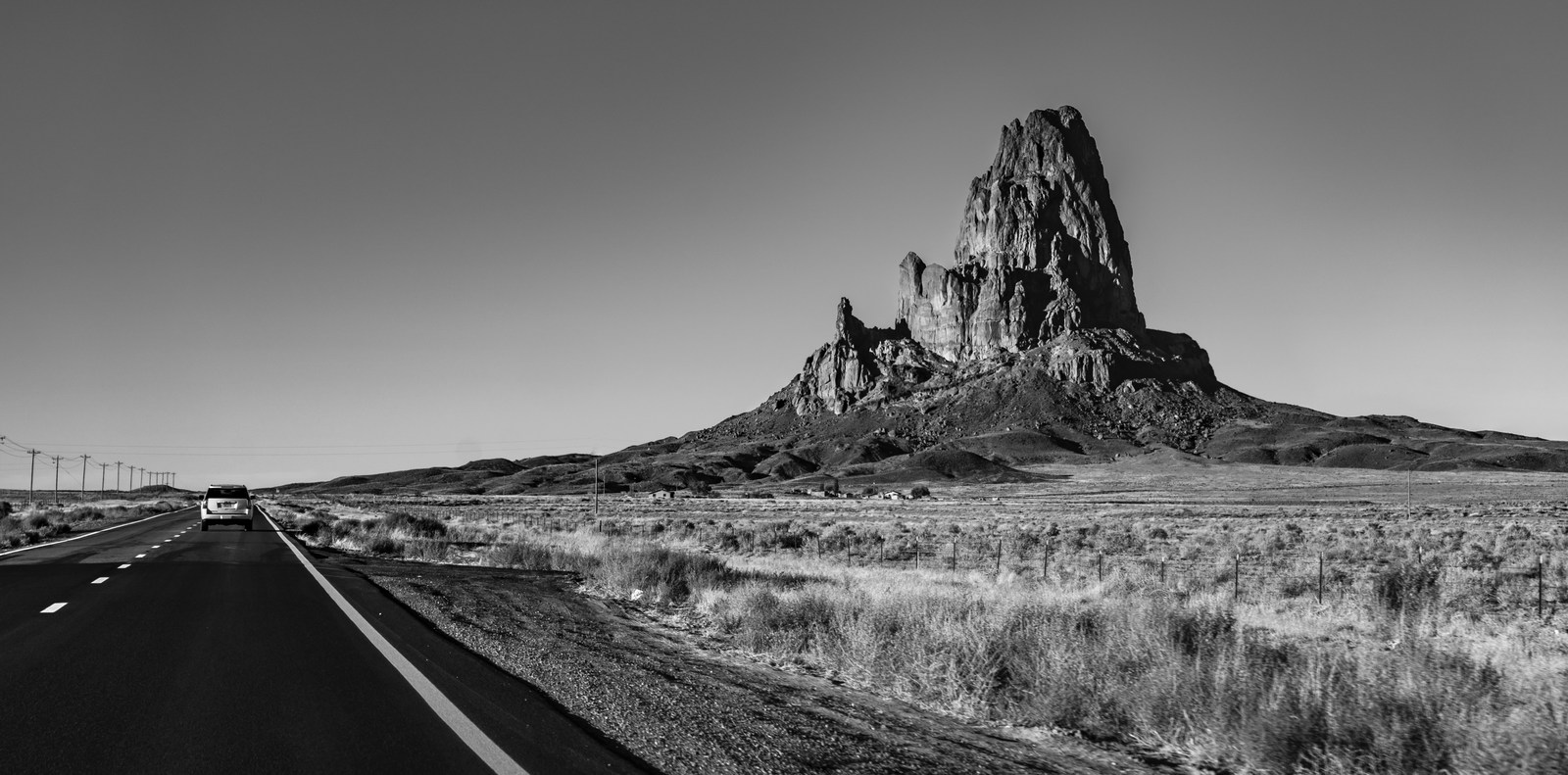 A road and a distant vehicle pass a solitary rock tower in monochrome.