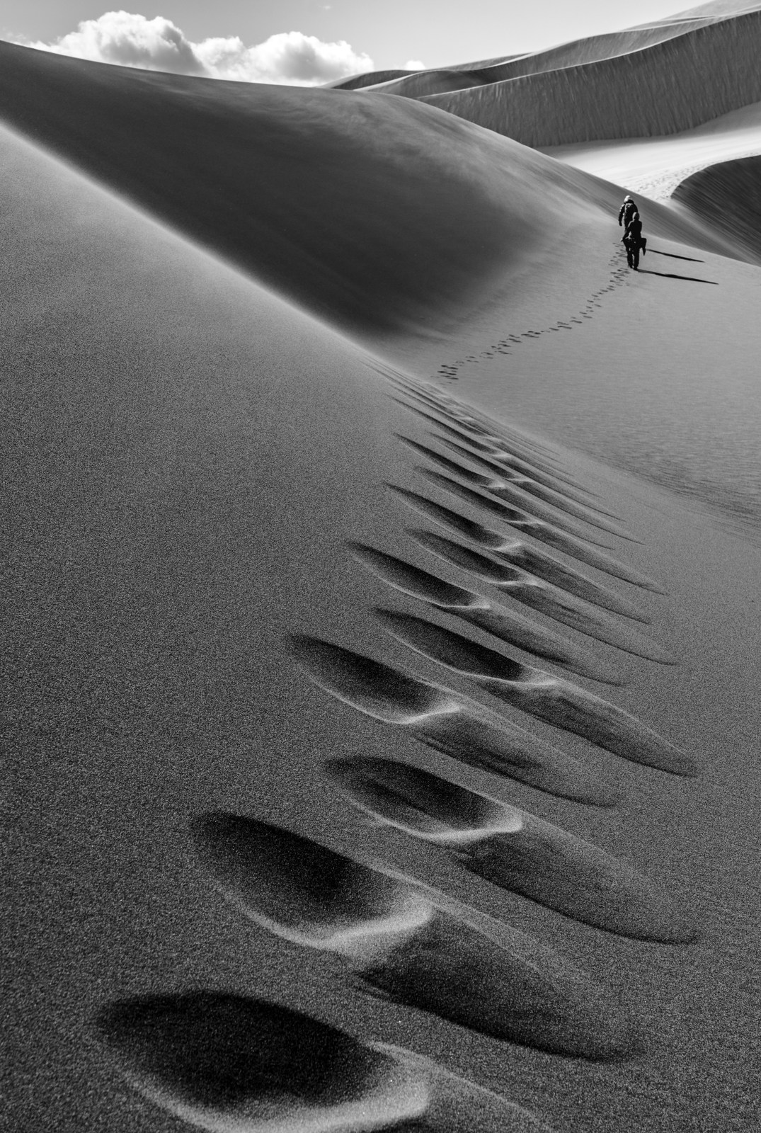 Footprints diminish along a steep dune toward two distant walkers.
