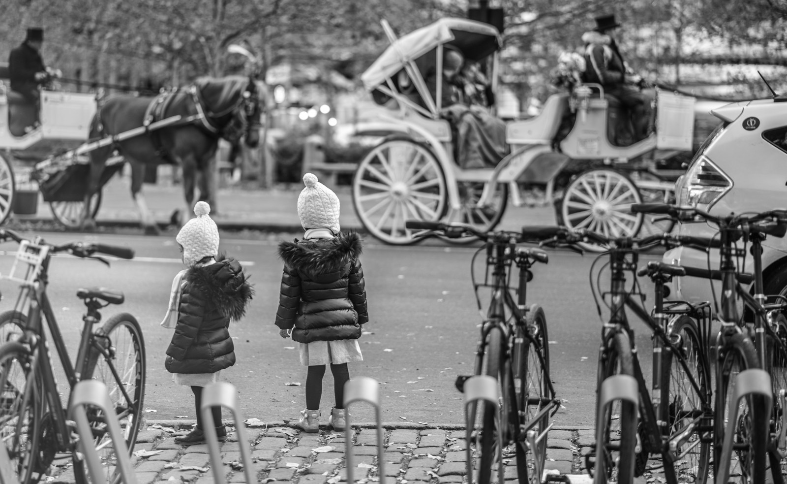 Two children in matching knit hats and dark coats watch horse carriages from between parked bicycles, in black and white.