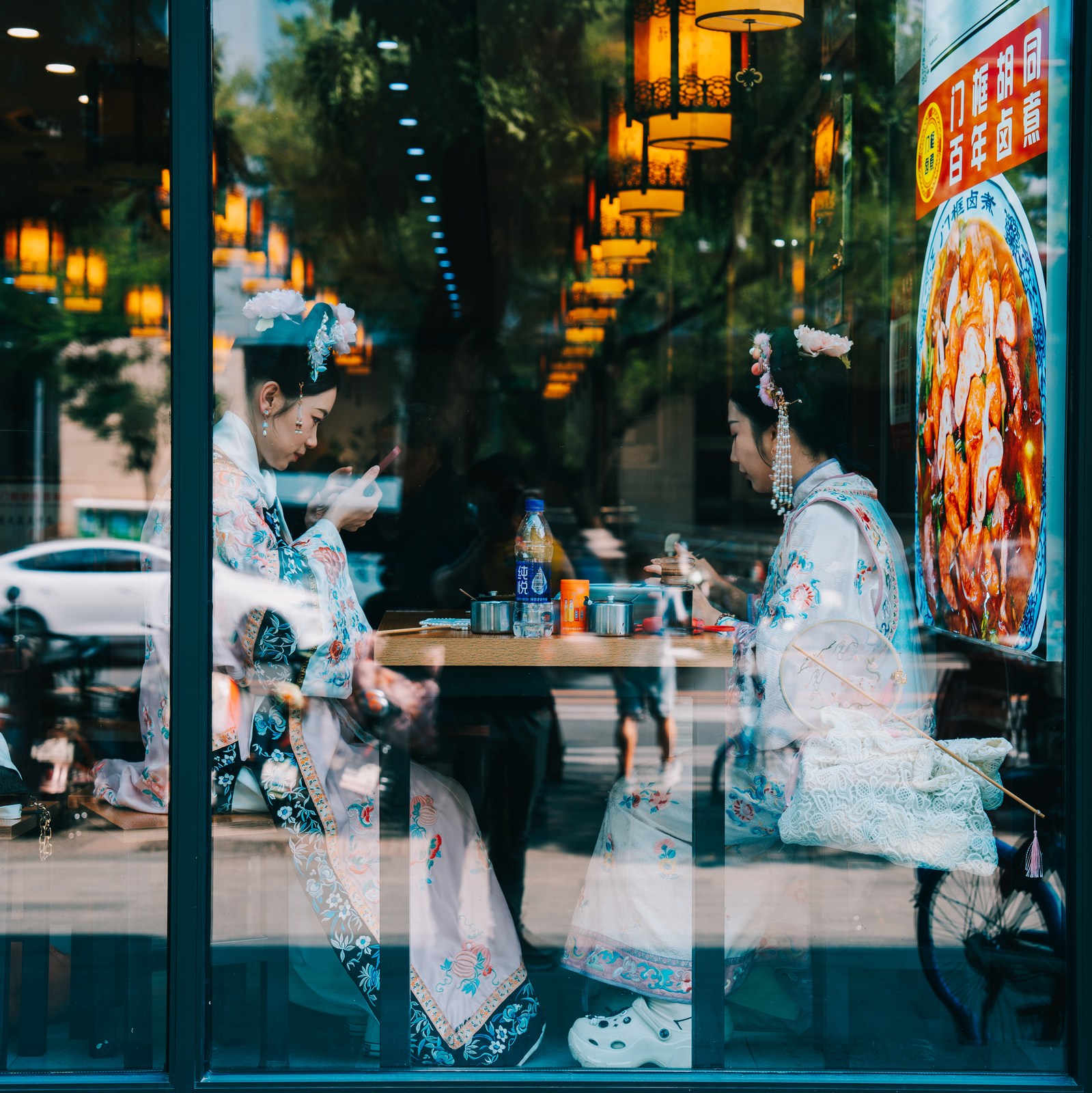 Two women sit across a restaurant table beneath lanterns, with street reflections crossing the window.