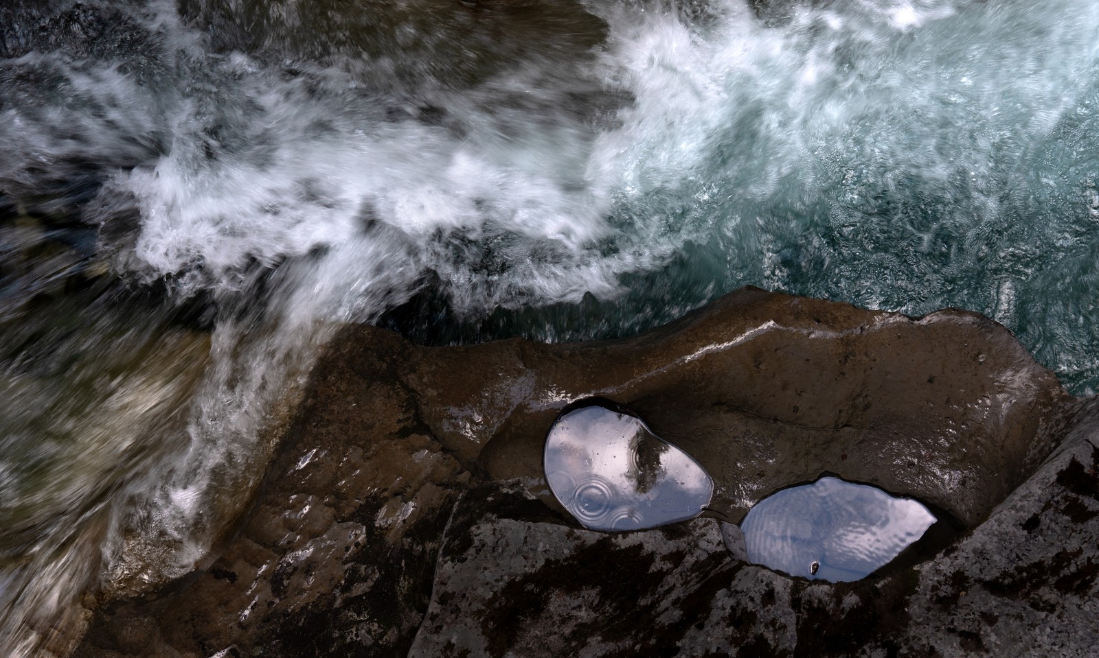 Two reflective pools in brown rock beside a rushing blue-white river.