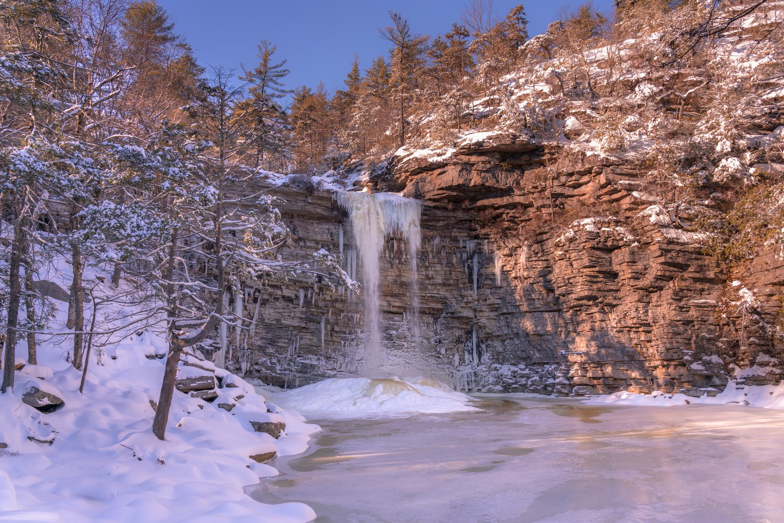 A waterfall drops over layered rock into an ice-covered pool surrounded by snow.