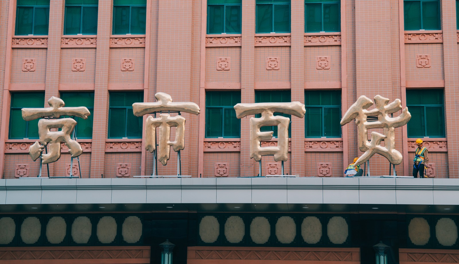 Two workers in yellow hard hats are beside oversized gold Chinese characters on a pink and green facade.